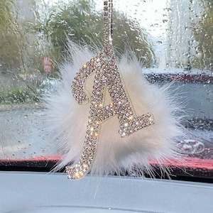 May include: A car rearview mirror hanging ornament featuring a large, silver-toned letter A encrusted with clear, sparkling rhinestones. The letter is centered on a fluffy, white faux fur pom-pom and suspended by a silver rhinestone chain.