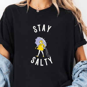 Könnte beinhalten: Schwarzes T-Shirt mit den weißen Worten "STAY SALTY". Das Shirt zeigt eine Grafik einer Person in einem gelben Regenmantel, die einen lila Regenschirm hält. Die Person geht im Regen. Das Shirt ist ein lässiger Stil.