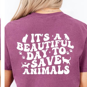 May include: Mauve t-shirt with a white veterinary symbol on the front and the phrase "It's a beautiful day to save animals" on the back. The design includes cat, paw print, and dog silhouettes.