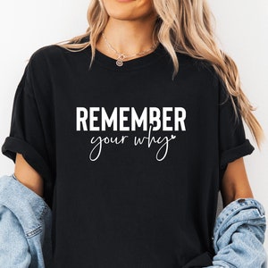 May include: Black t-shirt with the white text "REMEMBER your why". The shirt has a crew neck design. A denim jacket is worn over the shoulders.