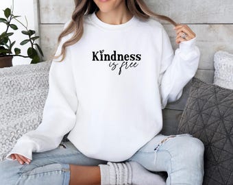 Könnte beinhalten: Ein weißes Sweatshirt mit dem schwarzen Schriftzug "Kindness is free". Das langärmlige Sweatshirt mit Rundhalsausschnitt wird von einer Person getragen, die blaue Jeans trägt. Das Bild ist vor einem Holzgrund aufgenommen.
