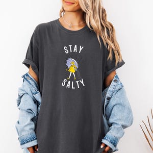 May include: Dark gray t-shirt with the words "STAY SALTY" in white. The graphic shows a person in a yellow raincoat and holding a purple umbrella in the rain. The shirt has rolled-up sleeves and a relaxed fit.