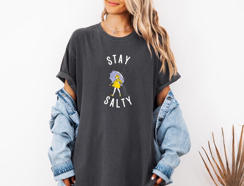 May include: Dark gray t-shirt with the words "STAY SALTY" in white. The graphic shows a person in a yellow raincoat and holding a purple umbrella in the rain. The shirt has rolled-up sleeves and a relaxed fit.