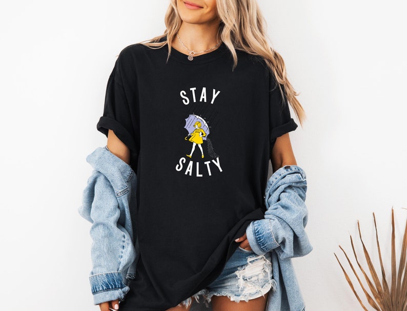 May include: Black t-shirt with the words "STAY SALTY" in white. The shirt features a graphic of a person in a yellow raincoat and holding a purple umbrella. The person is walking in the rain. The shirt is a casual style.