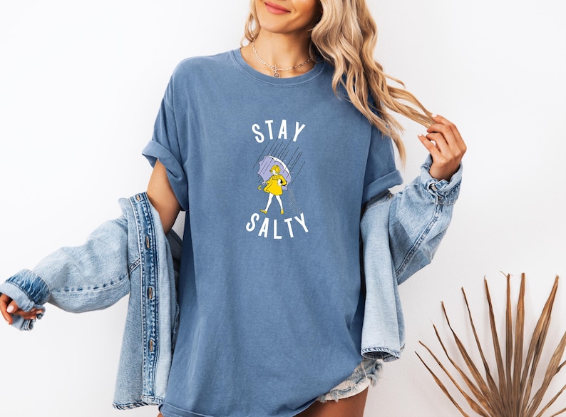 May include: A blue t-shirt with the words "STAY SALTY" and a graphic of a person with an umbrella in the rain. The person is wearing a yellow dress. The shirt is a casual style.