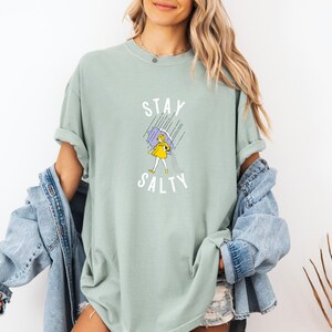 May include: Sage green t-shirt with the words "Stay Salty" above a cartoon illustration of a person in a yellow dress holding a purple umbrella in the rain. The shirt is a comfortable, oversized fit.