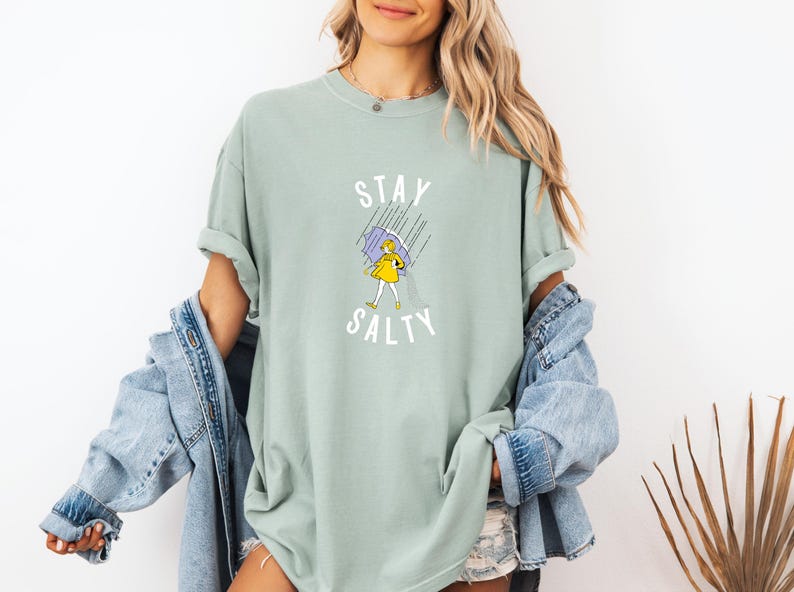 May include: Sage green t-shirt with the words "Stay Salty" above a cartoon illustration of a person in a yellow dress holding a purple umbrella in the rain. The shirt is a comfortable, oversized fit.