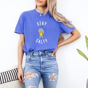 May include: A blue t-shirt with the words "STAY SALTY" in white, featuring a yellow cartoon character with an umbrella. The shirt is paired with blue jeans.
