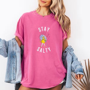 May include: A vibrant pink t-shirt with the words "STAY SALTY" in white, featuring a cartoon illustration of a person under an umbrella. The shirt has a relaxed fit and rolled-up sleeves.