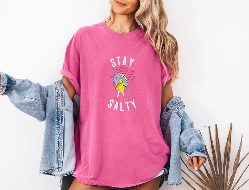 May include: A vibrant pink t-shirt with the words "STAY SALTY" in white, featuring a cartoon illustration of a person under an umbrella. The shirt has a relaxed fit and rolled-up sleeves.