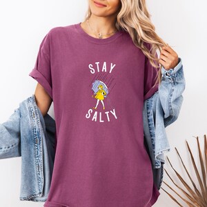 May include: A maroon t-shirt with the words "STAY SALTY" and a graphic of a person in a yellow dress holding an umbrella in the rain. The person is wearing a denim jacket.