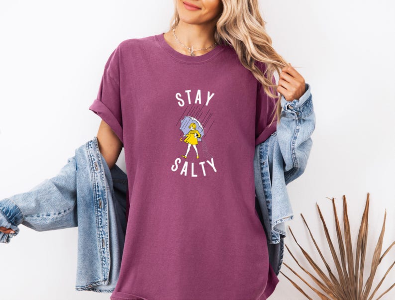 May include: A maroon t-shirt with the words "STAY SALTY" and a graphic of a person in a yellow dress holding an umbrella in the rain. The person is wearing a denim jacket.