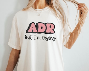 May include: Cream-colored t-shirt with the text "ADR" in pink bubble letters outlined in black. Below, the phrase "but I'm trying" is written in a simple black font. The shirt has short sleeves and a relaxed fit.