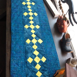 May include: A quilted table runner in shades of blue and yellow. The runner features a repeating pattern of yellow diamonds set against a blue background. The runner is rectangular and appears to be handmade.