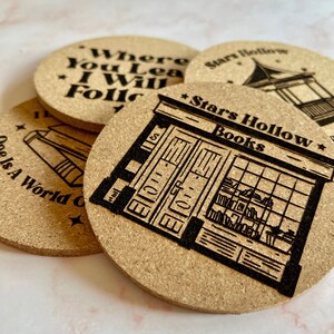 K&ouml;nnte beinhalten: Vier Korkuntersetzer mit schwarzen lasergravierten Designs. Ein Untersetzer zeigt den Text "Where You Lead I Will Follow". Ein weiterer Untersetzer zeigt den Text "Stars Hollow Books" und eine Ladenfront-Illustration. Der dritte Untersetzer zeigt den Text "Stars Hollow" und eine Illustration eines Pavillons. Der vierte Untersetzer zeigt den Text "One Is A World Of" und eine Illustration eines Buches.