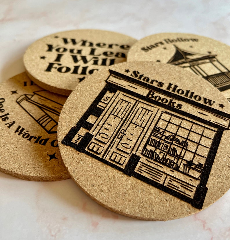 K&ouml;nnte beinhalten: Vier Korkuntersetzer mit schwarzen lasergravierten Designs. Ein Untersetzer zeigt den Text "Where You Lead I Will Follow". Ein weiterer Untersetzer zeigt den Text "Stars Hollow Books" und eine Ladenfront-Illustration. Der dritte Untersetzer zeigt den Text "Stars Hollow" und eine Illustration eines Pavillons. Der vierte Untersetzer zeigt den Text "One Is A World Of" und eine Illustration eines Buches.