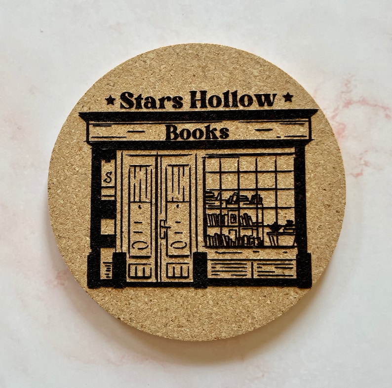 K&ouml;nnte beinhalten: Ein brauner Korkuntersetzer mit einer schwarz-wei&szlig;en Illustration einer Ladenfront mit dem Text "Stars Hollow Books" &uuml;ber der T&uuml;r.