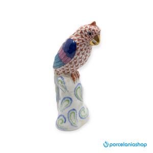 May include: A detailed porcelain parrot figurine perched on a white base with blue and green paisley designs. The parrot features a red crest, a gold beak, and a patterned body with shades of brown, blue, and pink. The figurine is a decorative item.