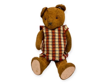Könnte beinhalten: Ein vintage brauner Teddybär, der eine rot-weiß karierte Weste trägt. Der Bär hat eine genähte Nase und schwarze Augen. Der Bär sitzt mit gekreuzten Beinen.