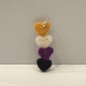 May include: A vertical pendant featuring four stacked felt hearts in yellow, white, purple, and black. The hearts are connected in a column, with a small silver-toned metal jump ring attached to the top yellow heart for hanging or attachment.