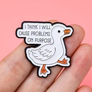 May include: White enamel pin featuring a cartoon goose with the text "I think I will cause problems on purpose".