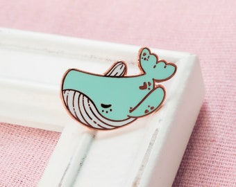 May include: A light blue whale enamel pin with a rose gold finish. The whale has a closed eye and a small heart on its tail fin.