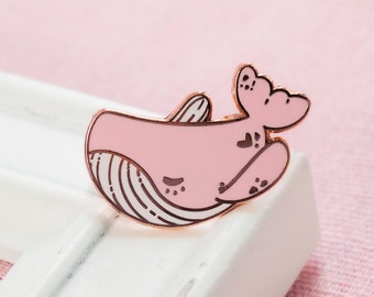 Puede incluir: Un pin de esmalte de oro rosa de una ballena rosa con vientre blanco y aleta caudal blanca. La ballena está enroscada con su aleta caudal sobre su cabeza.