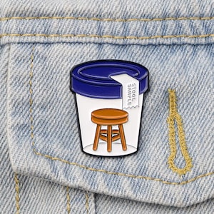 May include: A blue and white enamel pin shaped like a small plastic container with a brown stool inside. The container has a white label that says "Stool Sample".
