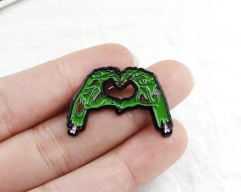 May include: Green zombie hands forming a heart shape. The hands are black with green details and pink accents. The pin is a fun and spooky accessory.