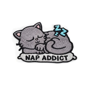 May include: Embroidered patch featuring a gray cat sleeping on a pillow. The cat has closed eyes and pink inner ears. The pillow has the words "NAP ADDICT" in black text. Blue "Z"s indicate the cat is sleeping.