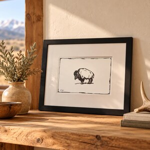 May include: A black and white ink drawing of a bison centered within a white mat and a black rectangular frame. The artwork features a signature in the bottom left corner and is displayed on a wooden surface next to a ceramic vase.