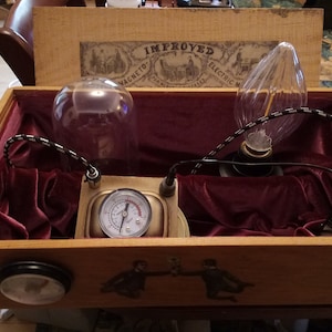 May include: A wooden box with a red velvet interior, containing a steampunk-style device with a glass dome, a light bulb, and a pressure gauge. The box lid features the text "IMPROVED MAGNETO ELECTRIC MACHINE".