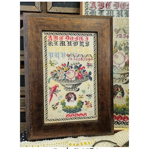 May include: A framed cross-stitch sampler with a floral design and the text "A B C D O F C J K L M N O R S" and "1234567890". The sampler is in a brown wooden frame with a decorative border. The title "Guth R 1874" and "A Reproduction" are printed below the frame.