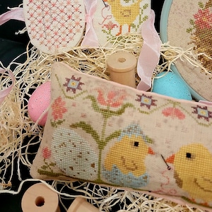 May include: A cross-stitch pillow with two chicks in a nest, surrounded by flowers and a pink ribbon. There are also two wooden spools and three cross-stitch eggs.