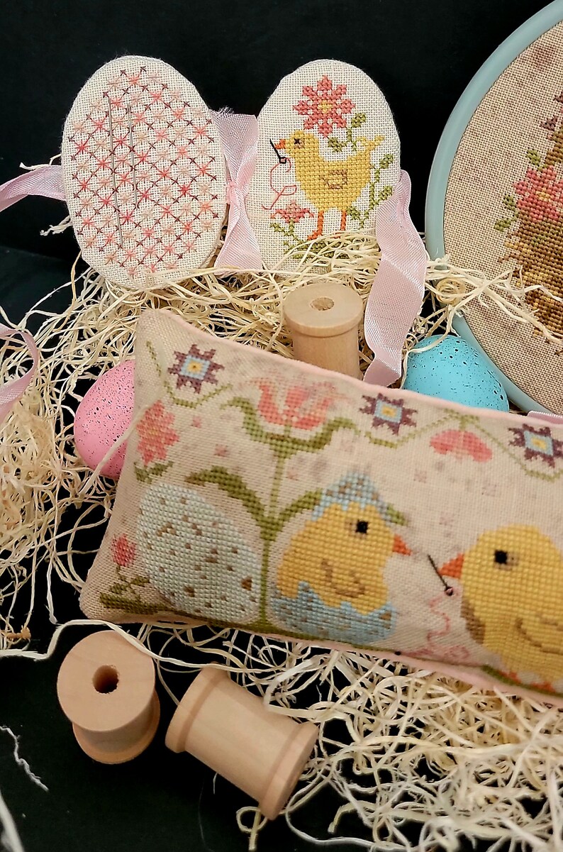 May include: A cross-stitch pillow with two chicks in a nest, surrounded by flowers and a pink ribbon. There are also two wooden spools and three cross-stitch eggs.