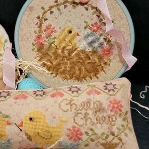 May include: Two cross-stitch embroidery hoops with a pastel pink and blue color scheme. The larger hoop features a nest with two yellow chicks and the text "Cheep Cheep". The smaller hoop features a single yellow chick with a needle and thread.