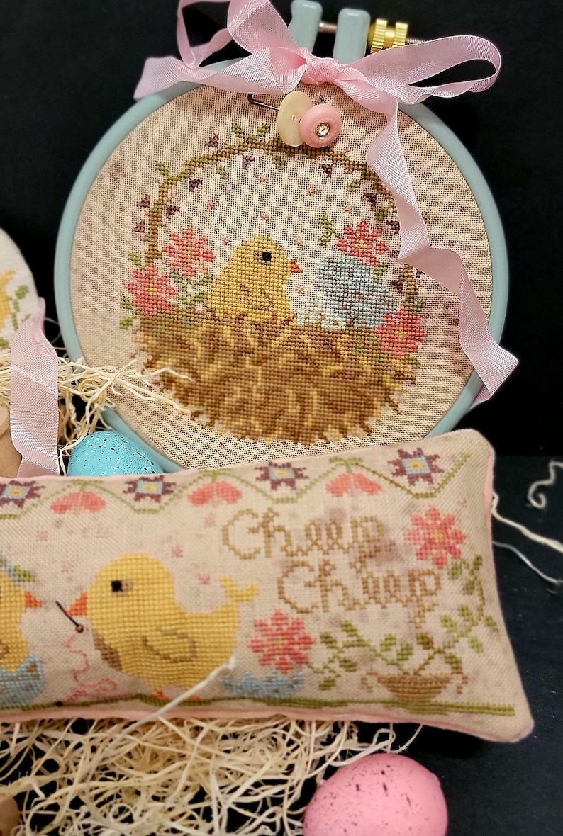 May include: Two cross-stitch embroidery hoops with a pastel pink and blue color scheme. The larger hoop features a nest with two yellow chicks and the text "Cheep Cheep". The smaller hoop features a single yellow chick with a needle and thread.