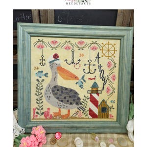 May include: Cross-stitch artwork titled "Captain Patchy Pelican" in a teal frame. The design features a pelican wearing a sailor hat, a lighthouse, anchors, and floral accents. The color palette includes shades of blue, orange, and beige.