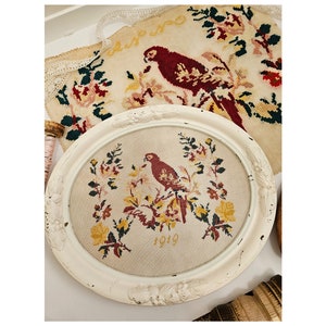 May include: A white oval frame with a cream-colored background featuring a cross-stitch design of a red bird perched on a branch with yellow and pink flowers. The design is dated "1919" and the frame has a white, ornate border. The text "Dutch Bird 1919" is written below the frame.