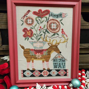 May include: A framed cross-stitch design featuring a reindeer pulling a wagon with a flower pot and the text "Jingle All The Way". The reindeer is wearing red and white striped stockings. The design is surrounded by a red frame.