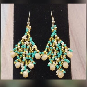 May include: A pair of dangle earrings with a teardrop shape. The earrings are made with turquoise, white, and gold beads. The earrings have a silver hook.