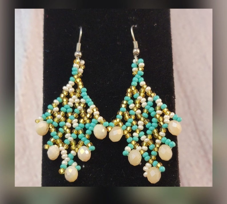May include: A pair of dangle earrings with a teardrop shape. The earrings are made with turquoise, white, and gold beads. The earrings have a silver hook.