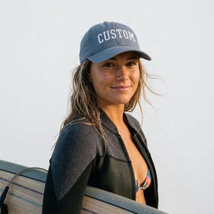 May include: A slate blue baseball cap featuring the word CUSTOM embroidered in white capital letters on the front panel. The cap has a structured crown, a curved brim, and a fabric strap closure at the back, providing a classic athletic look.