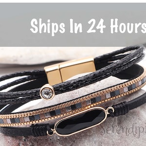 May include: A multi-layered black leather bracelet with gold accents and a black oval charm. The bracelet features a small clear rhinestone and the word "Serendipity" is visible in the image.
