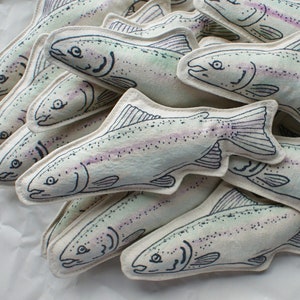 May include: A group of 10 handmade plush trout fish toys. The fish are made of white fabric with a light blue and pink watercolor design. The fish have black embroidered details and are arranged in a pile on a white surface.