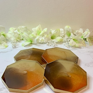 May include: Set of four hexagonal resin coasters with gold-colored edges. The coasters feature a gradient of brown, amber, and gold tones. The coasters are arranged on a white marble surface, with white flowers in the background.
