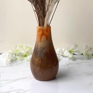 May include: A decorative vase with a gradient of colors, transitioning from orange to brown. The vase holds dried flowers and grasses in shades of brown and cream. White flowers are in the background.