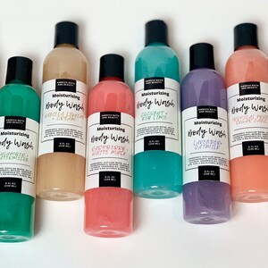 May include: Eight clear plastic bottles of body wash with black lids and white labels. The labels have the text "Moisturizing Body Wash" and the scent of each body wash. The scents are:  "Watermelon", "Vanilla & Coconut Cream", "Raspberry White Musk", "Coconut & Key Lime", "Lavender Oatmilk", "Tropical Fruit Sugar Cake", "Milk & Honey".