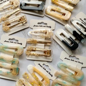 May include: Assortment of hair clips in various colors and designs. The clips include rectangular and barrette styles, with gold-tone metal hardware. Some clips feature glitter, while others have marbled or solid color patterns. The packaging includes the text "Amreva Bath and Beauty".
