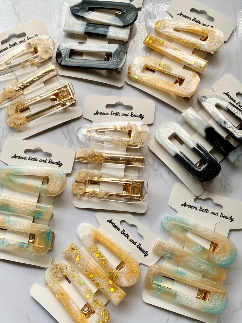 May include: Assortment of hair clips in various colors and designs. The clips include rectangular and barrette styles, with gold-tone metal hardware. Some clips feature glitter, while others have marbled or solid color patterns. The packaging includes the text "Amreva Bath and Beauty".